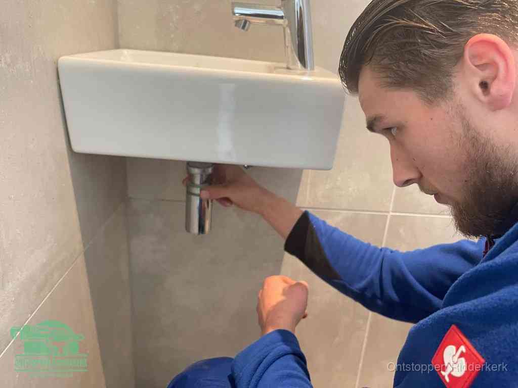 Een monteur draait met de hand de zwanenhalssifon onder een fonteinwasbak in de toiletruimte los om de bron van rioollucht in Ridderkerk te vinden