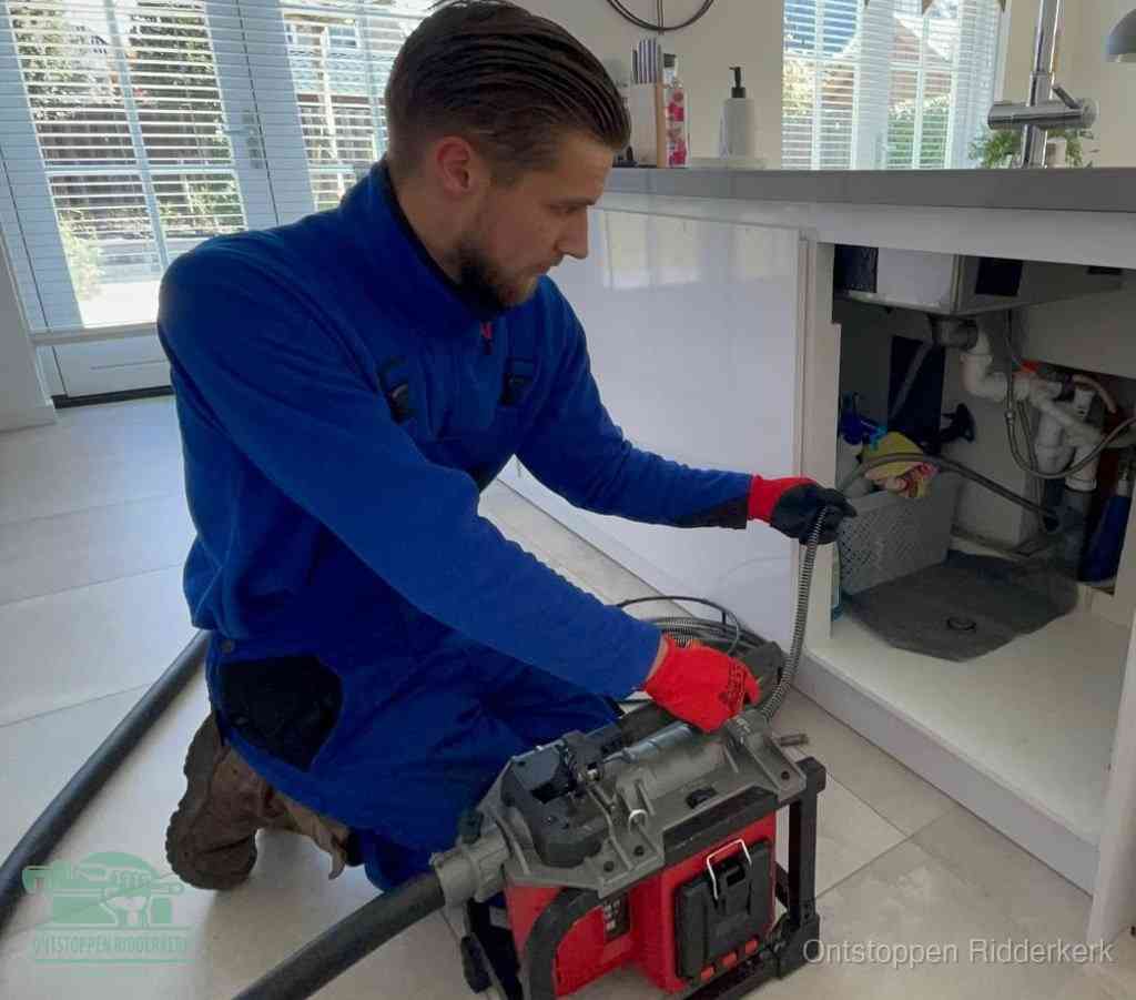 Een monteur met rode handschoenen knielt in een keuken en voert een elektrische ontstoppingsveer in tijdens het professioneel afvoer ontstoppen in Ridderkerk